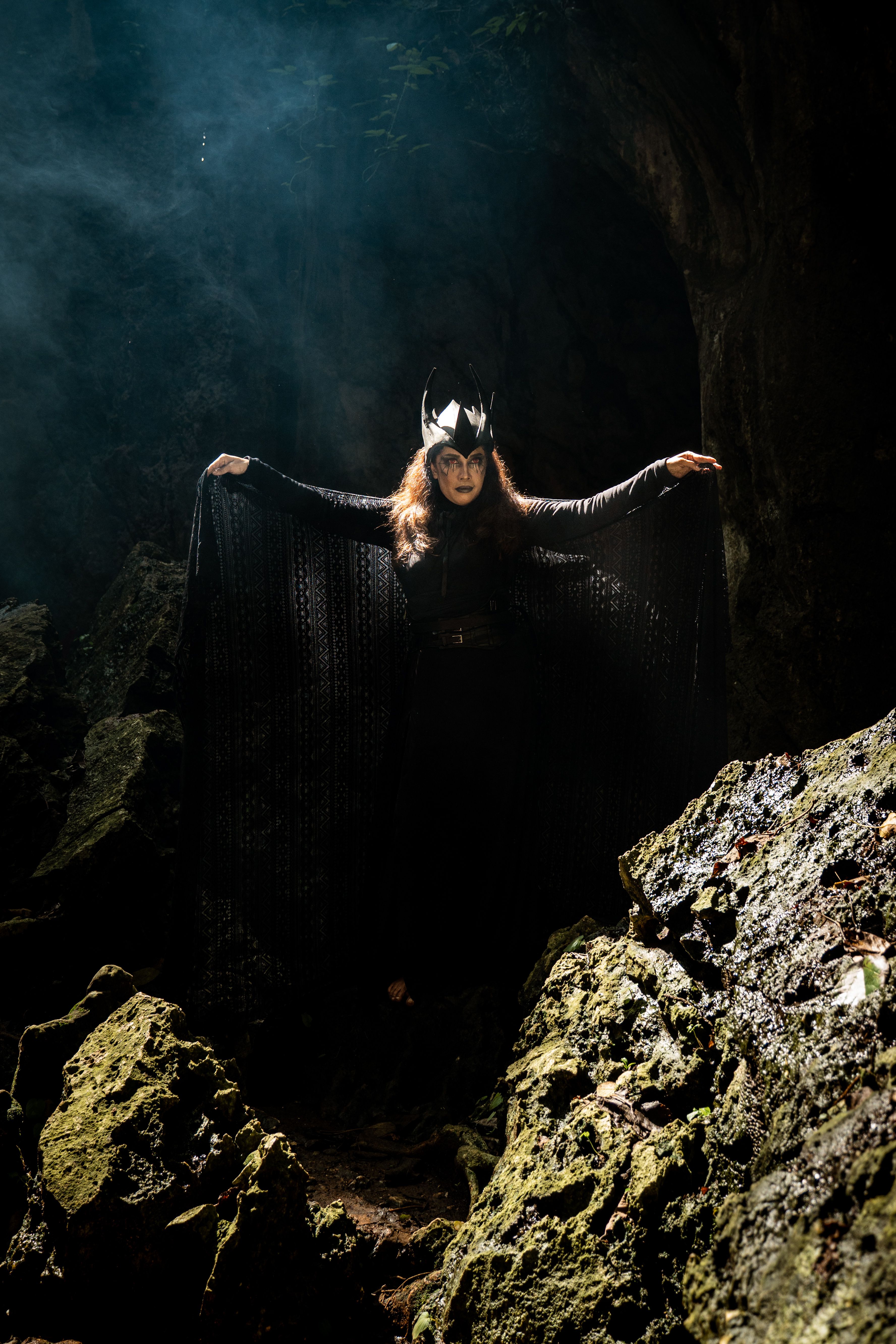A woman in black with mystical attire stands in a forest cave, creating an enigm