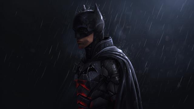 Batman stands in profile clad in a dark tactical suit as rain falls heavily around him at night.