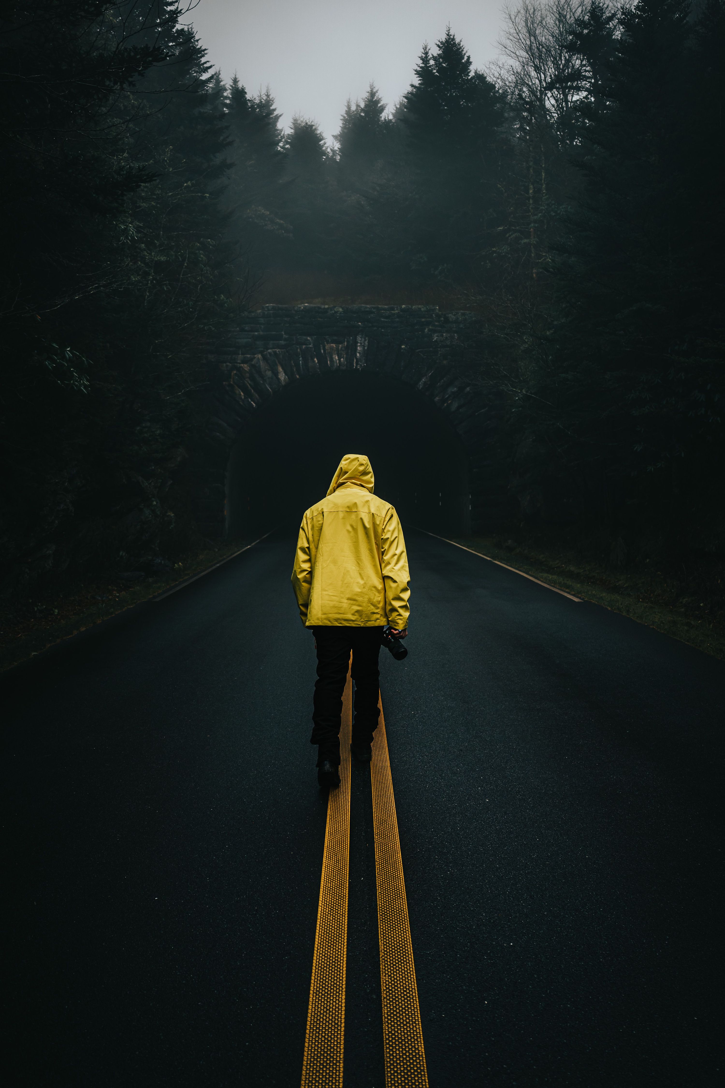 A solitary figure in a yellow jacket walks down a foggy road towards a dark tunn