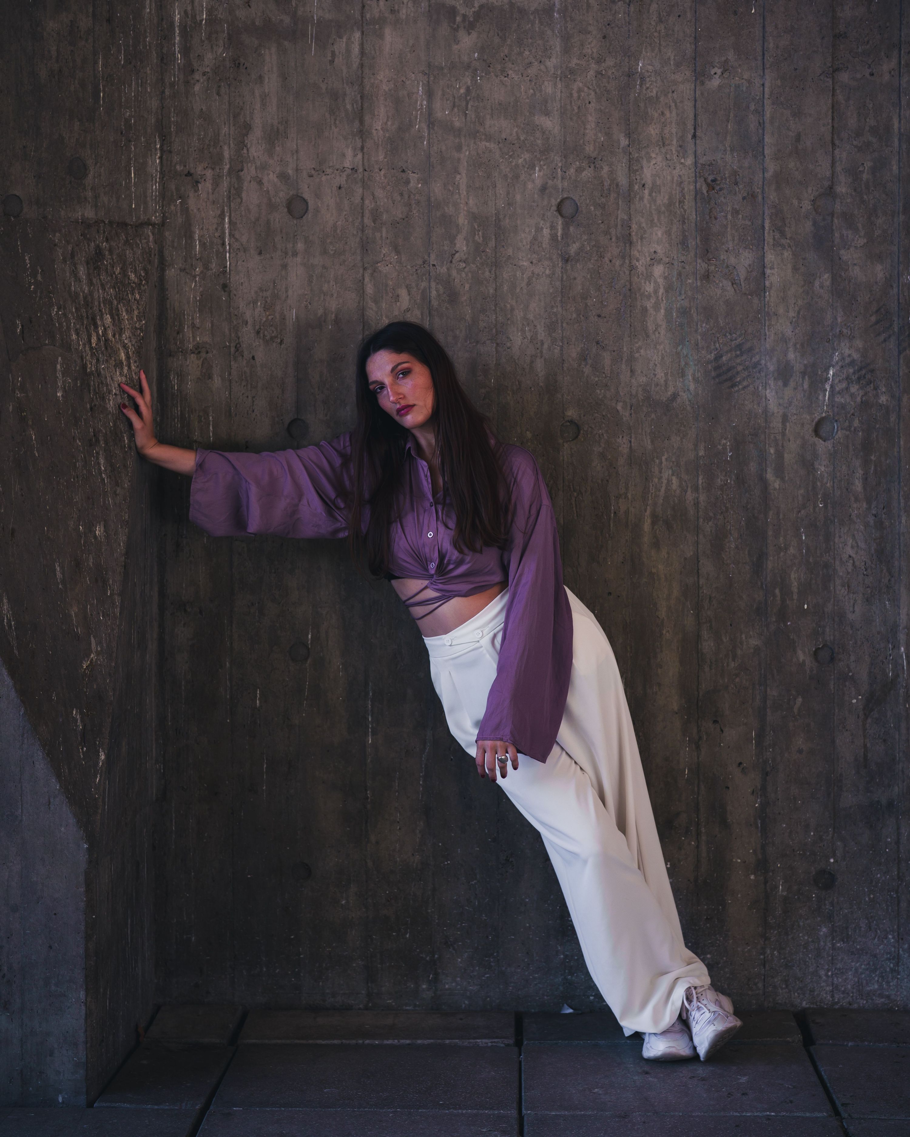 Fashion model in purple top and white pants leans dramatically on a concrete wal