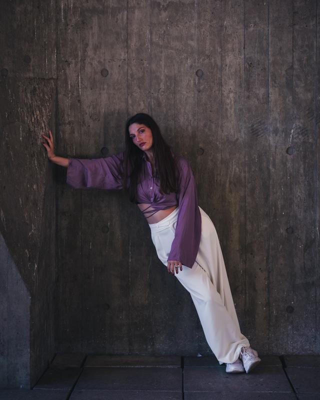 Fashion model in purple top and white pants leans dramatically on a concrete wal